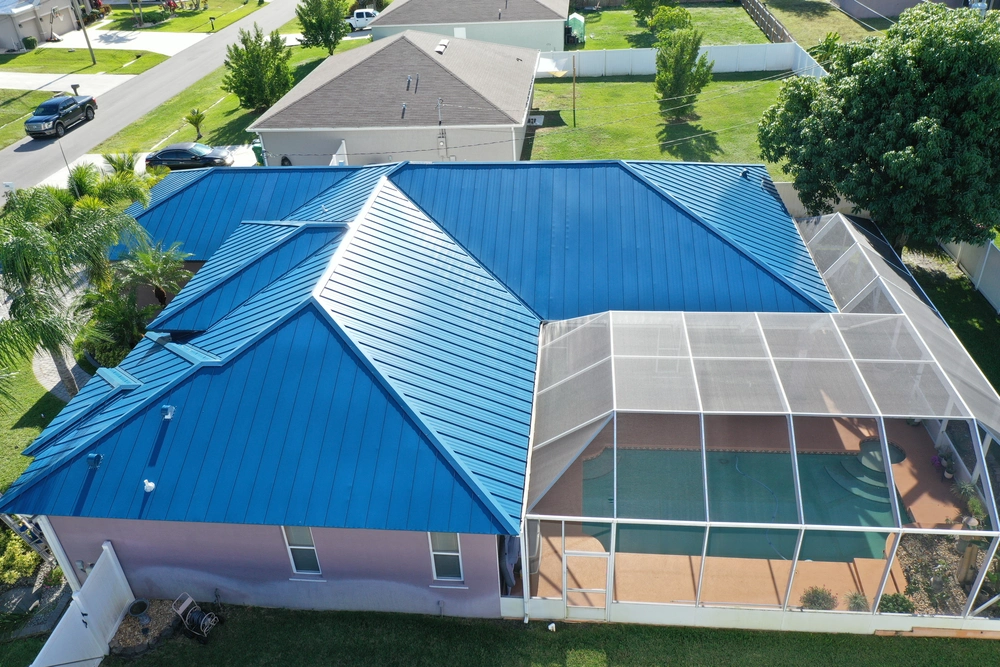 Aerial view of a house with a bright blue metal roof. The backyard features a screened-in pool area enclosed with a mesh structure. Surrounding houses and green lawns are visible, along with trees and a parked car on the street.
