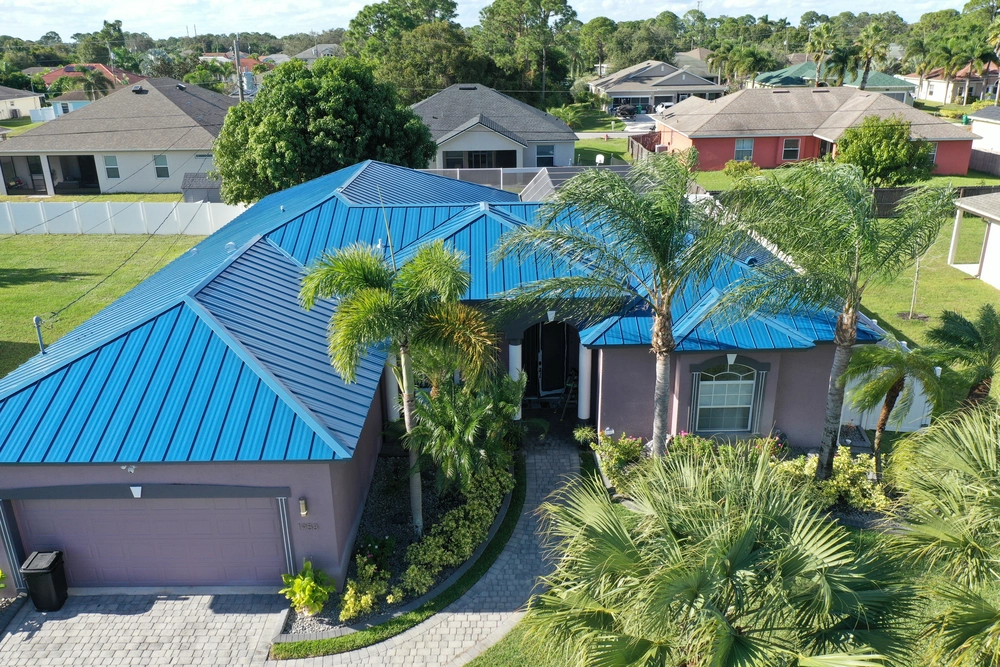 Aerial view of a residential house with a blue metal roof surrounded by palm trees and a well-maintained lawn. The neighborhood features similar houses with different colored roofs, set against a backdrop of trees and a partly cloudy sky.
