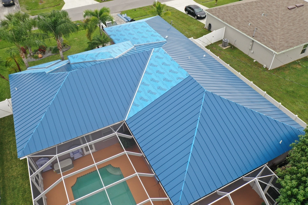 Aerial view of a house with blue metal roofing. Part of the roof has blue underlayment visible where tiles are missing. The house features a screened patio with a swimming pool, surrounded by grass and other houses.