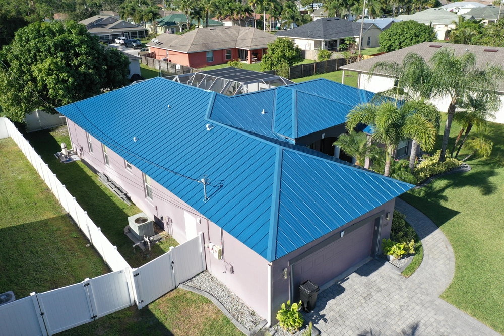 Aerial view of a residential property featuring a house with a bright blue metal roof. The house is surrounded by a white fence and has a small yard with palm trees. Neighboring homes and lush greenery are visible in the background.