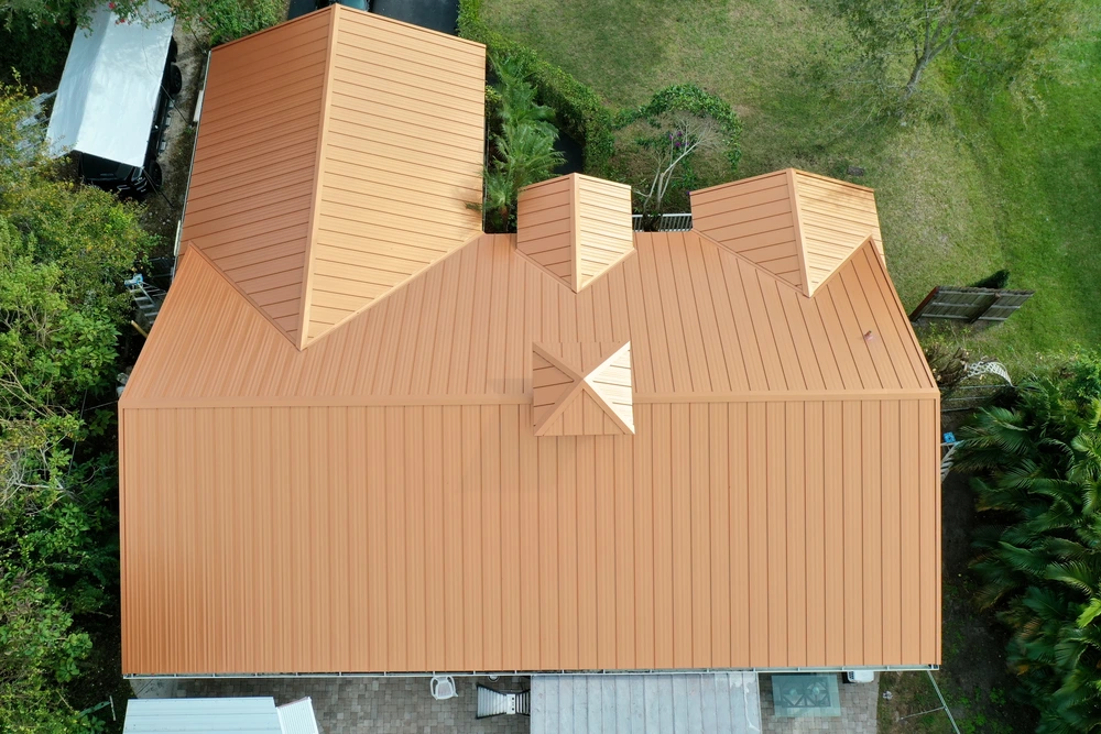 Aerial view of a building with an orange metal roof featuring multiple peaks and ridges, surrounded by greenery and concrete areas.