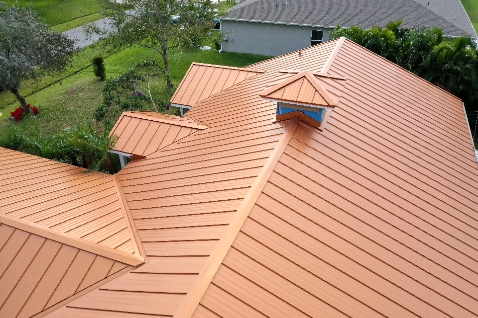 Aerial view of a modern house with a copper-colored metal roof featuring several dormer windows. The roof's surface is smooth and shiny. Surrounding the house are trees, a neatly trimmed lawn, and a neighboring building.