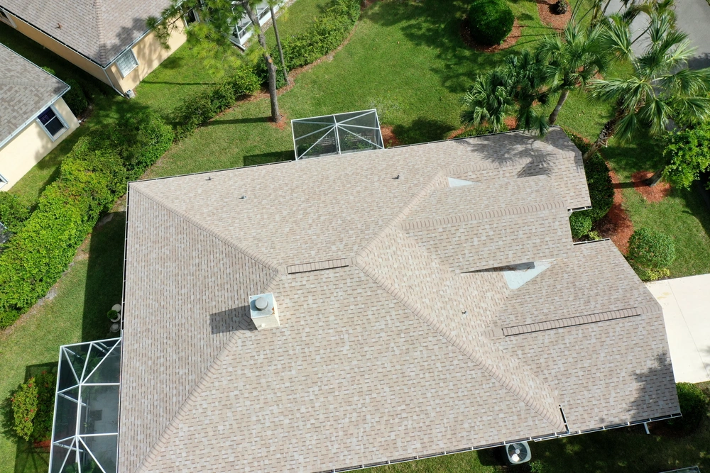 Aerial view of a house with a beige shingled roof, featuring a central chimney and skylights. The roof is surrounded by a green lawn, bushes, and trees. Part of a driveway is visible to the right.