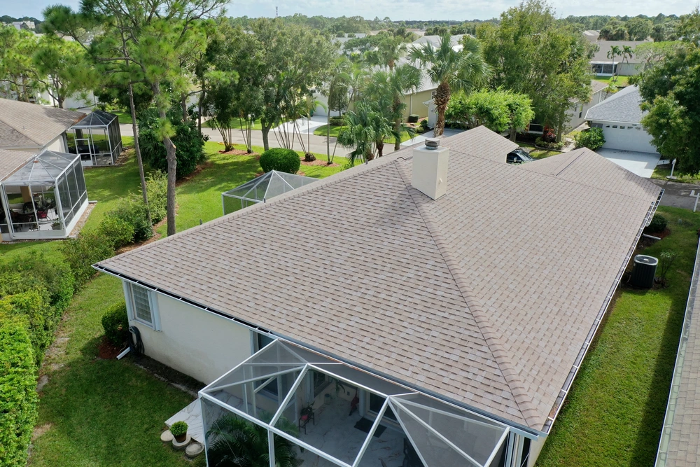 Aerial view of a suburban house with a brown shingle roof. The backyard features screened enclosures. Trees and other houses are visible in the background on a sunny day.