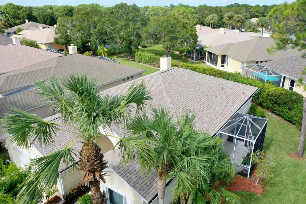 Aerial view of suburban homes with tan roofs and screened patios, surrounded by lush greenery and palm trees. The neighborhood has a peaceful, tropical atmosphere with well-maintained landscaping and dense trees in the background.