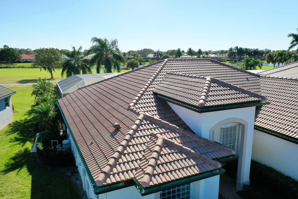 Aerial view of a single-story house with a red-tiled roof. The property is surrounded by green lawns and palm trees, set against a clear blue sky. The house has a modern design with several roof sections and white walls with dark trim.