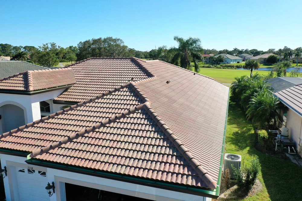 Aerial view of a house with a terracotta-tiled roof surrounded by greenery. Nearby are other homes and a golf course. The sky is clear and blue, suggesting a sunny day.
