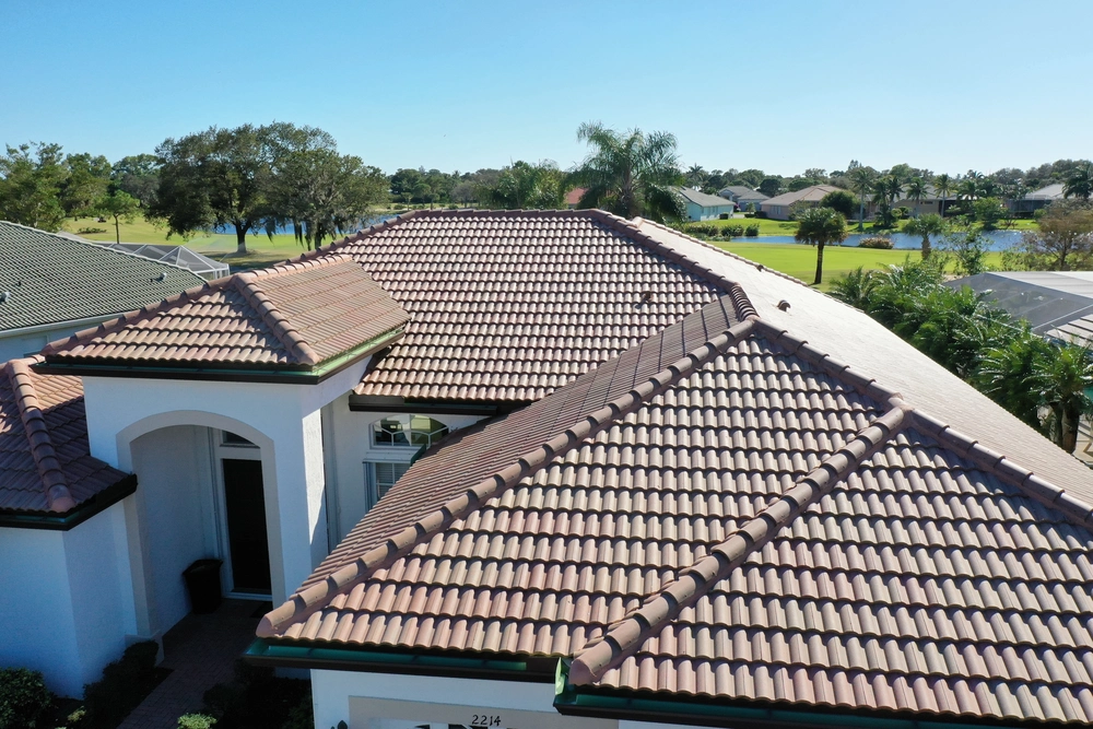 Aerial view of a house with a tiled roof, featuring multiple peaks and angles. The roof tiles are a mix of brown and terracotta shades. Greenery and other homes are visible in the background under a clear blue sky.