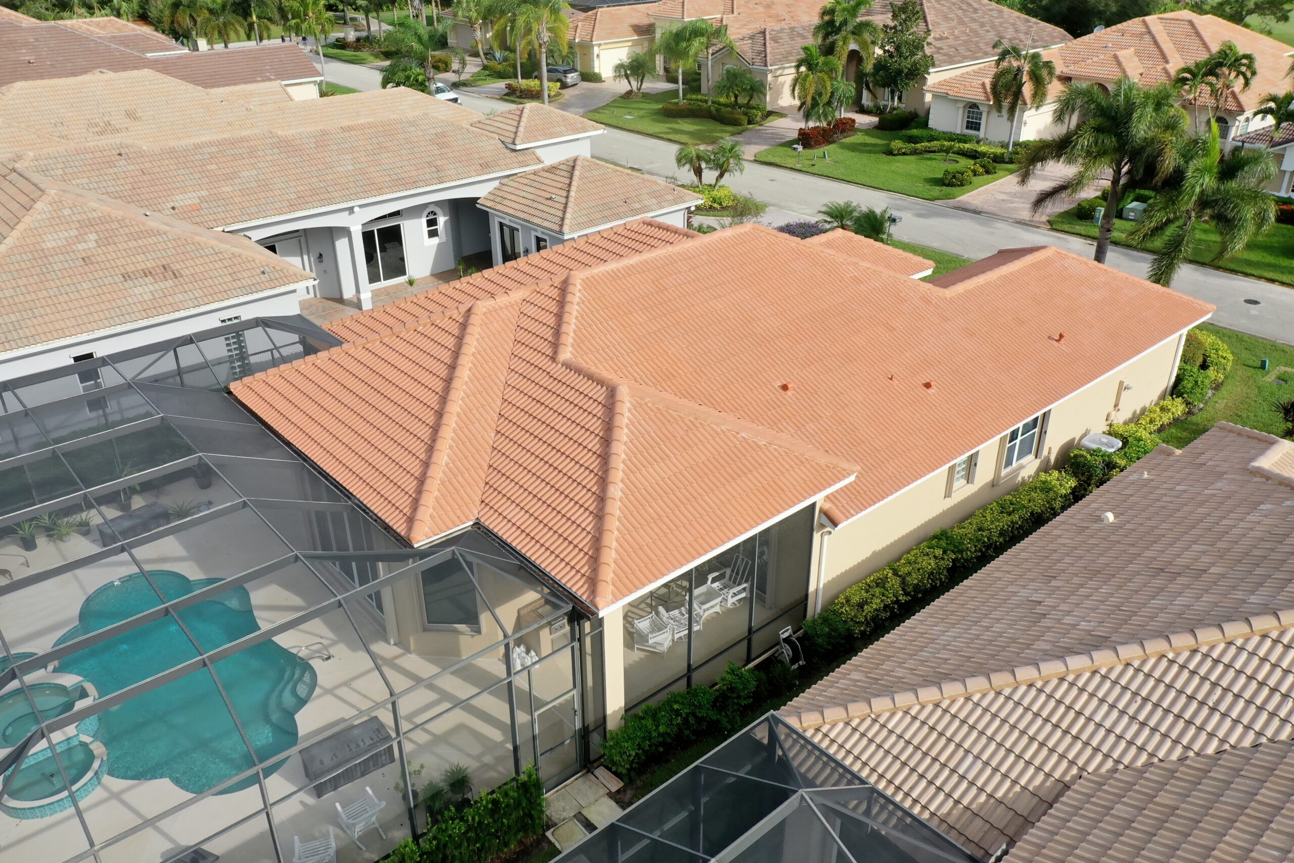 Aerial view of a suburban neighborhood in Port Saint Lucie with tan, concrete tile roof houses. One home stands out with its screened-in pool area and patio. The uniquely shaped pool is surrounded by lush greenery, while nearby streets are lined with palm trees and other charming homes in the Caramel Estate.