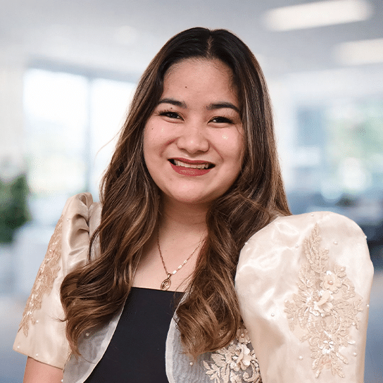 A woman with long wavy hair, wearing a traditional embroidered beige top with puffed sleeves and a black inner dress, smiles at the camera. The background is a bright, blurred indoor office space.