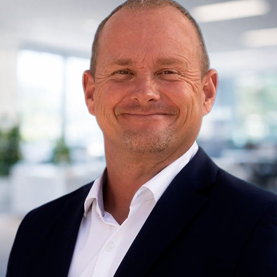 A middle-aged man with short hair, wearing a dark suit jacket and white shirt, smiles at the camera in a bright, modern office setting.