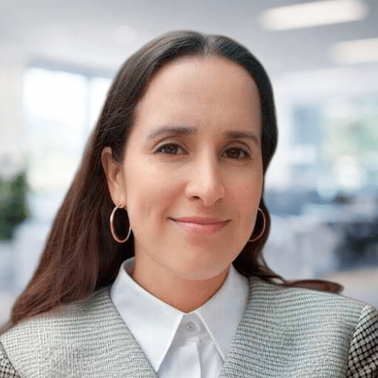 A woman with long dark hair, wearing a gray blazer, white collared shirt, and hoop earrings, smiles softly in a bright, blurred office setting.