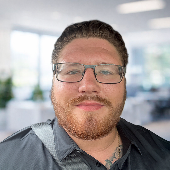 A man with short brown hair, a beard, and glasses is wearing a black collared shirt and a shoulder strap. He is smiling and standing in a bright, blurred office setting.
